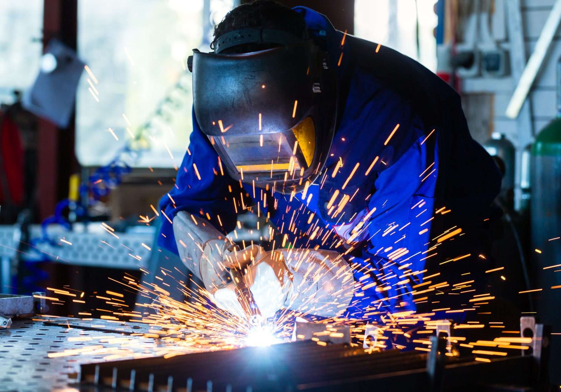 Welder bonding metal with welding device in workshop, lots of sparks to be seen, he wears welding googles