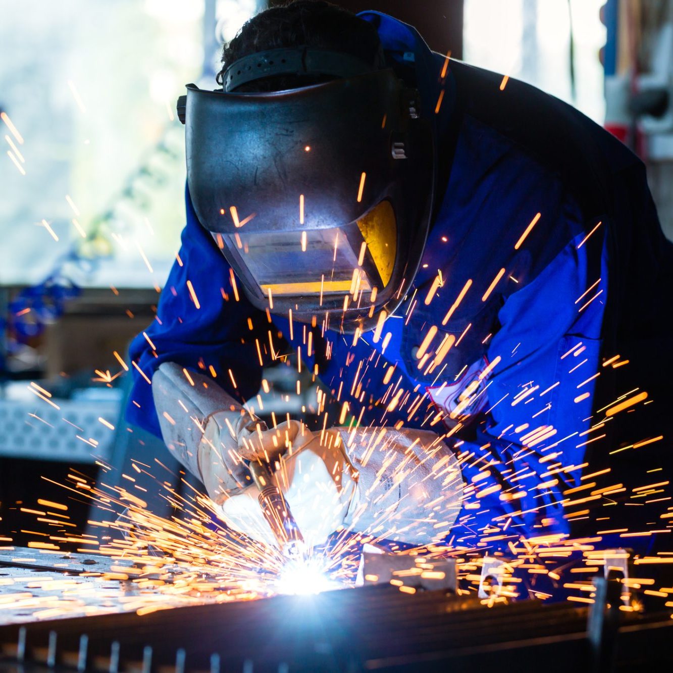 Welder bonding metal with welding device in workshop, lots of sparks to be seen, he wears welding googles